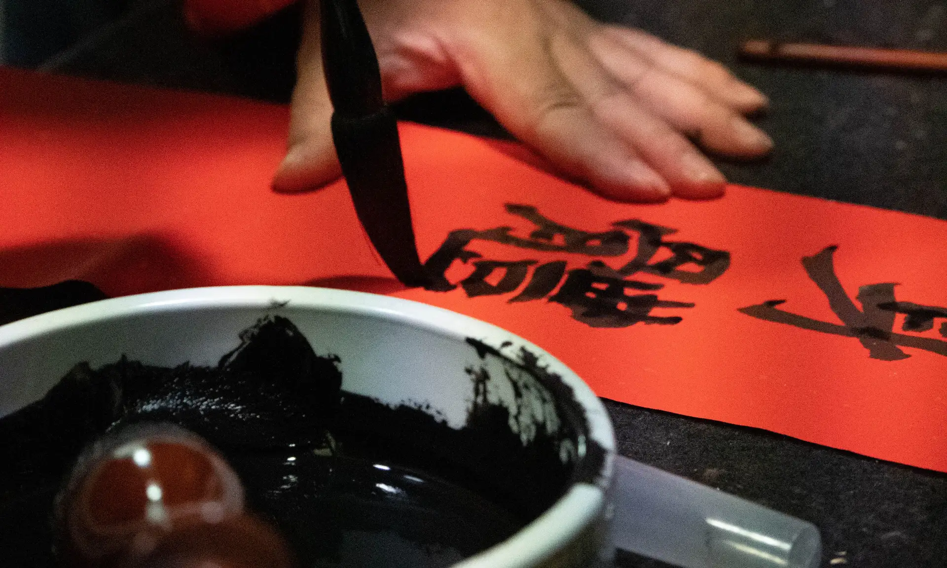 A calligrapher's brush touching red paper mid-stroke, forming bold Chinese characters in black ink.